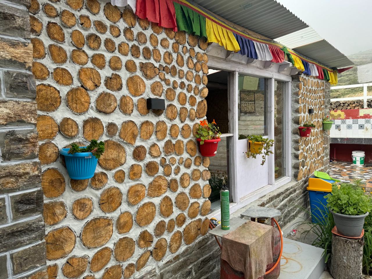 Stone wall of Karma Inn lined with stacked firewood, potted flowers and prayer flags